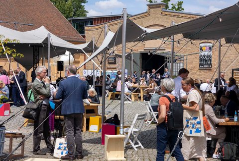 Eine Fotografie zeigt einen belebten Innenhof mit temporären Zeltdächern, Holzbänken und Tischen, zahlreiche Menschen sitzen, stehen oder kommen miteinander ins Gespräch, während historische Backsteinbauten den Rahmen bilden, wodurch das Bild eine offene Veranstaltungsatmosphäre vermittelt und für Austausch, Begegnung und Baukultur als gesellschaftliches Ereignis steht.