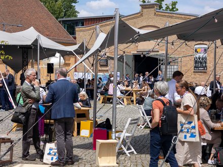 Eine Fotografie zeigt einen belebten Innenhof mit temporären Zeltdächern, Holzbänken und Tischen, zahlreiche Menschen sitzen, stehen oder kommen miteinander ins Gespräch, während historische Backsteinbauten den Rahmen bilden, wodurch das Bild eine offene Veranstaltungsatmosphäre vermittelt und für Austausch, Begegnung und Baukultur als gesellschaftliches Ereignis steht.