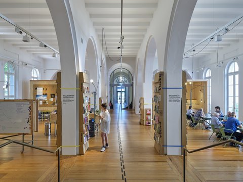 Großer heller Raum mit hohen Rundbögen, Holzfußboden und mehreren Sitzgruppen, in der Mitte steht eine Person vor einem Bücherregal