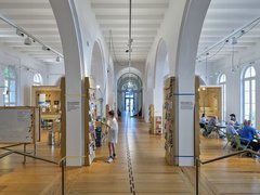 Großer heller Raum mit hohen Rundbögen, Holzfußboden und mehreren Sitzgruppen, in der Mitte steht eine Person vor einem Bücherregal