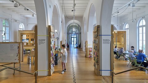 Großer heller Raum mit hohen Rundbögen, Holzfußboden und mehreren Sitzgruppen, in der Mitte steht eine Person vor einem Bücherregal
