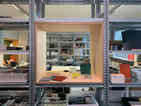 „Blick durch ein industrielles Metallregal auf einen Holzkasten mit verschiedenen Material- und Farbmustern in einer Mustersammlung.“