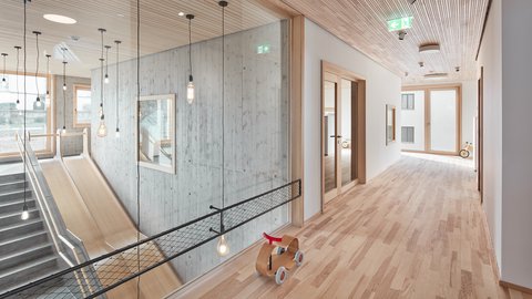 Heller Flur mit Holzdecke und -boden, links Treppe mit Rutsche hinter Glaswand, rechts offene Türen, auf dem Boden ein kleines Holzfahrzeug