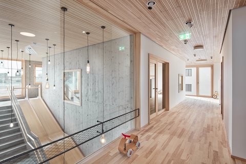 Heller Flur mit Holzdecke und -boden, links Treppe mit Rutsche hinter Glaswand, rechts offene Türen, auf dem Boden ein kleines Holzfahrzeug