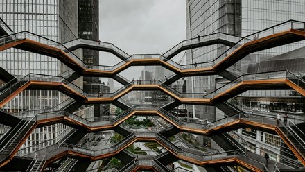  Große, wabenförmige Treppenanlage mit mehreren Ebenen zwischen hohen Glasgebäuden; wenige Personen auf den Stegen; urbanes Umfeld, bedeckter Himmel; Blick von innen nach außen.