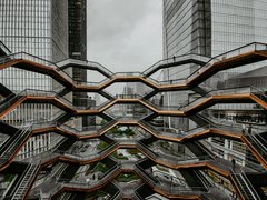  Große, wabenförmige Treppenanlage mit mehreren Ebenen zwischen hohen Glasgebäuden; wenige Personen auf den Stegen; urbanes Umfeld, bedeckter Himmel; Blick von innen nach außen.