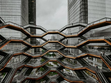  Große, wabenförmige Treppenanlage mit mehreren Ebenen zwischen hohen Glasgebäuden; wenige Personen auf den Stegen; urbanes Umfeld, bedeckter Himmel; Blick von innen nach außen.