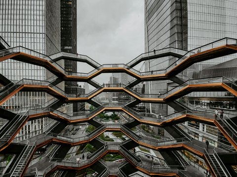  Große, wabenförmige Treppenanlage mit mehreren Ebenen zwischen hohen Glasgebäuden; wenige Personen auf den Stegen; urbanes Umfeld, bedeckter Himmel; Blick von innen nach außen.