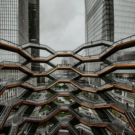  Große, wabenförmige Treppenanlage mit mehreren Ebenen zwischen hohen Glasgebäuden; wenige Personen auf den Stegen; urbanes Umfeld, bedeckter Himmel; Blick von innen nach außen.