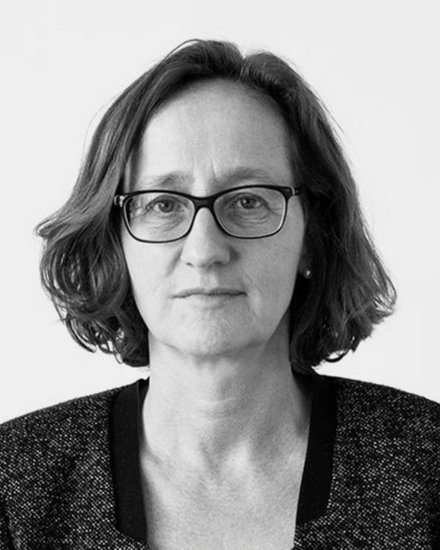 "Schwarz-Weiß-Porträtfoto einer Frau mittleren Alters mit schulterlangem, welligem dunklem Haar. Sie trägt eine rechteckige Brille mit dunklem Gestell und einen dunklen Blazer mit strukturiertem Stoff über einem hellen Oberteil. Sie blickt direkt in die Kamera mit neutralem, nachdenklichem Gesichtsausdruck. Der Hintergrund ist neutral und hell. Das Foto hat einen professionellen Charakter."