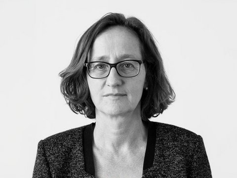 "Schwarz-Weiß-Porträtfoto einer Frau mittleren Alters mit schulterlangem, welligem dunklem Haar. Sie trägt eine rechteckige Brille mit dunklem Gestell und einen dunklen Blazer mit strukturiertem Stoff über einem hellen Oberteil. Sie blickt direkt in die Kamera mit neutralem, nachdenklichem Gesichtsausdruck. Der Hintergrund ist neutral und hell. Das Foto hat einen professionellen Charakter."
