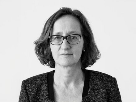 "Schwarz-Weiß-Porträtfoto einer Frau mittleren Alters mit schulterlangem, welligem dunklem Haar. Sie trägt eine rechteckige Brille mit dunklem Gestell und einen dunklen Blazer mit strukturiertem Stoff über einem hellen Oberteil. Sie blickt direkt in die Kamera mit neutralem, nachdenklichem Gesichtsausdruck. Der Hintergrund ist neutral und hell. Das Foto hat einen professionellen Charakter."