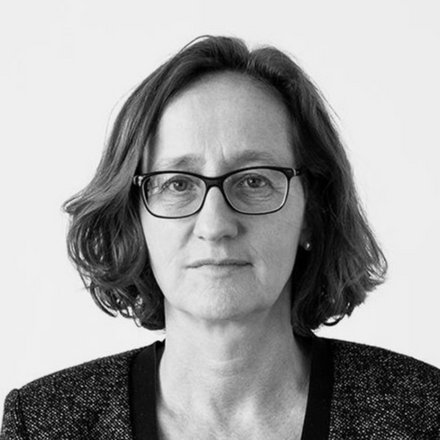 "Schwarz-Weiß-Porträtfoto einer Frau mittleren Alters mit schulterlangem, welligem dunklem Haar. Sie trägt eine rechteckige Brille mit dunklem Gestell und einen dunklen Blazer mit strukturiertem Stoff über einem hellen Oberteil. Sie blickt direkt in die Kamera mit neutralem, nachdenklichem Gesichtsausdruck. Der Hintergrund ist neutral und hell. Das Foto hat einen professionellen Charakter."