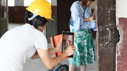  Person mit gelbem Schutzhelm dokumentiert eine Wand mit einem Tablet; im Hintergrund schreibt eine weitere Person Notizen.