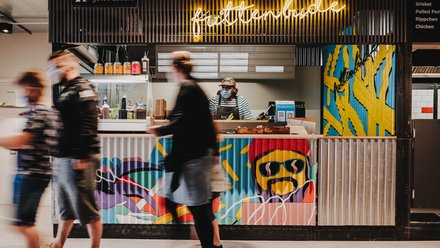 Essensstand mit leuchtender Beschriftung, Bedienung hinter der Theke und vorbeigehenden Gästen.