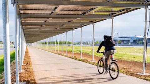 Überdachter Rad- und Fußweg unter einer modernen Photovoltaikanlage; eine Person fährt mit dem Fahrrad über den Weg.