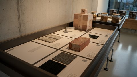  Ausstellungstisch mit beschrifteten Tafeln und Materialproben: Ziegelstein, massiver Holzblock, Kachel‑ und Textilproben sowie ein Metallbeschlag; hinten Gläser mit Erdproben.