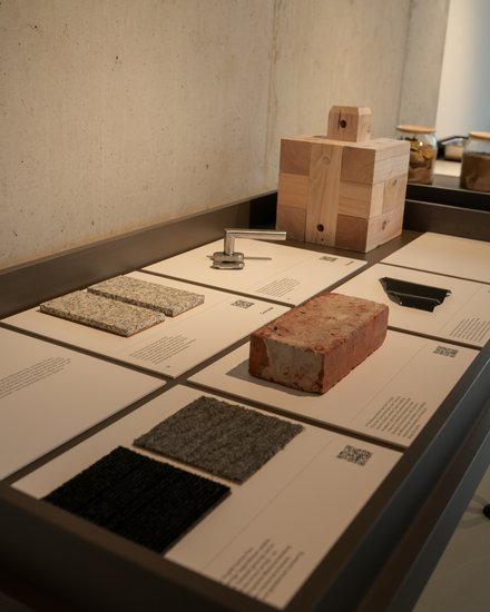  Ausstellungstisch mit beschrifteten Tafeln und Materialproben: Ziegelstein, massiver Holzblock, Kachel‑ und Textilproben sowie ein Metallbeschlag; hinten Gläser mit Erdproben.