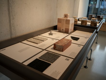  Ausstellungstisch mit beschrifteten Tafeln und Materialproben: Ziegelstein, massiver Holzblock, Kachel‑ und Textilproben sowie ein Metallbeschlag; hinten Gläser mit Erdproben.