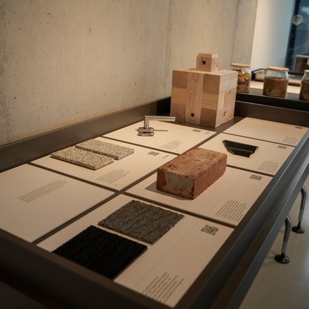  Ausstellungstisch mit beschrifteten Tafeln und Materialproben: Ziegelstein, massiver Holzblock, Kachel‑ und Textilproben sowie ein Metallbeschlag; hinten Gläser mit Erdproben.