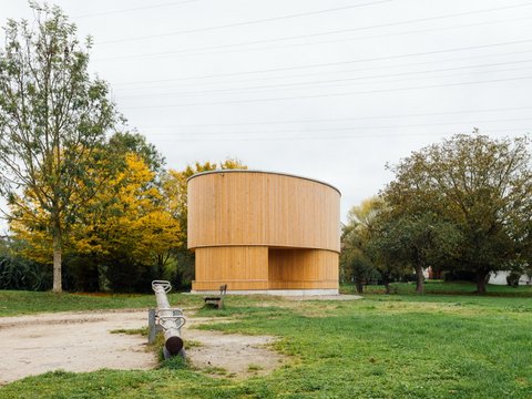 "Modernes zylindrisches Holzgebäude mit vertikalem Lamellenfassade in hellem Naturton. Das Gebäude steht auf einer grünen Wiese umgeben von alten Laubbäumen. Links ein metallischer Wasserpumpen-Hydranten. Rechts herbstlich gefärbte Bäume in Gelb und Braun. Bewölkter Himmel."