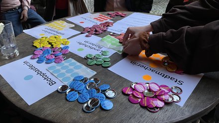 Runder Tisch mit farbigen Buttons und Klebepunkten; Hände sortieren die Abzeichen. Auf Zetteln liegen Kategorien, u. a. „Stadtmacher:in“.