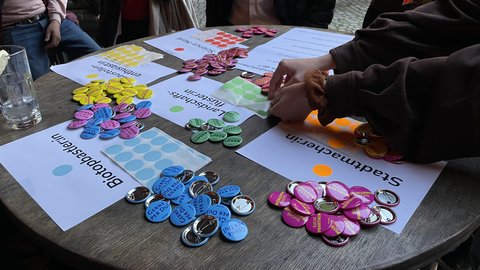 Runder Tisch mit farbigen Buttons und Klebepunkten; Hände sortieren die Abzeichen. Auf Zetteln liegen Kategorien, u. a. „Stadtmacher:in“.