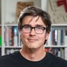 Porträt vor Bücherregal: Person mit dunklem, mittellangem Haar und schwarzer Brille, schwarzes T‑Shirt, blickt freundlich in die Kamera.