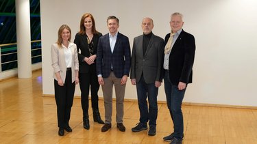 Sechs erwachsene Personen stehen nebeneinander in einem hellen Innenraum mit Holzfußboden und neutraler Wand, sie tragen überwiegend formelle bis halbformelle Kleidung und blicken lächelnd in die Kamera, was das Bild als offizielles Gruppenfoto eines Leitungsgremiums oder einer verantwortlichen Arbeitsgruppe kennzeichnet und Zusammenhalt, Zusammenarbeit und institutionelle Verantwortung sichtbar macht.