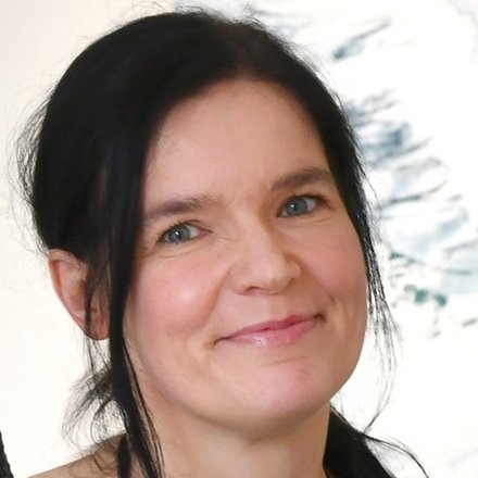 Porträtfoto derselben Frau in Farbe. Sie hat dunkelbraunes Haar, das teilweise zur Seite fällt, blaue Augen und ein warmherziges Lächeln. Sie trägt ein schwarzes Oberteil mit offenem Ausschnitt. Der unscharfe Hintergrund deutet auf eine Büroumgebung hin. Die Aufnahme wirkt professionell und zugänglich."