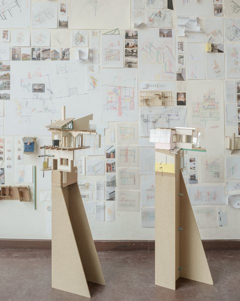Eine perspektivische Aufnahme eines Ateliers oder Ausstellungsraums, in dem zwei hohe Architekturmodelle auf Sperrholzsockeln vor einer mit Zeichnungen bedeckten Wand stehen. Das linke Modell zeigt eine komplexe, fragmentarische Gebäudestruktur mit Holzkonstruktionen, einem Kamin und einer grünen Dachstütze. Das rechte Modell hat eine rosa Wand und große Fenster. Die gesamte Rückwand ist dicht mit architektonischen Skizzen, Plänen, kleinen Fotos und gefalteten Papiermodellen bedeckt. Die Fülle an Studien vermittelt den Eindruck eines intensiven kreativen Prozesses. Der Boden ist aus dunklem Holz.