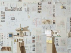 Eine perspektivische Aufnahme eines Ateliers oder Ausstellungsraums, in dem zwei hohe Architekturmodelle auf Sperrholzsockeln vor einer mit Zeichnungen bedeckten Wand stehen. Das linke Modell zeigt eine komplexe, fragmentarische Gebäudestruktur mit Holzkonstruktionen, einem Kamin und einer grünen Dachstütze. Das rechte Modell hat eine rosa Wand und große Fenster. Die gesamte Rückwand ist dicht mit architektonischen Skizzen, Plänen, kleinen Fotos und gefalteten Papiermodellen bedeckt. Die Fülle an Studien vermittelt den Eindruck eines intensiven kreativen Prozesses. Der Boden ist aus dunklem Holz.