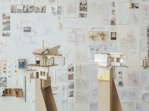 Eine perspektivische Aufnahme eines Ateliers oder Ausstellungsraums, in dem zwei hohe Architekturmodelle auf Sperrholzsockeln vor einer mit Zeichnungen bedeckten Wand stehen. Das linke Modell zeigt eine komplexe, fragmentarische Gebäudestruktur mit Holzkonstruktionen, einem Kamin und einer grünen Dachstütze. Das rechte Modell hat eine rosa Wand und große Fenster. Die gesamte Rückwand ist dicht mit architektonischen Skizzen, Plänen, kleinen Fotos und gefalteten Papiermodellen bedeckt. Die Fülle an Studien vermittelt den Eindruck eines intensiven kreativen Prozesses. Der Boden ist aus dunklem Holz.