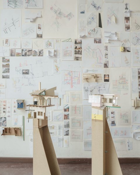 Eine perspektivische Aufnahme eines Ateliers oder Ausstellungsraums, in dem zwei hohe Architekturmodelle auf Sperrholzsockeln vor einer mit Zeichnungen bedeckten Wand stehen. Das linke Modell zeigt eine komplexe, fragmentarische Gebäudestruktur mit Holzkonstruktionen, einem Kamin und einer grünen Dachstütze. Das rechte Modell hat eine rosa Wand und große Fenster. Die gesamte Rückwand ist dicht mit architektonischen Skizzen, Plänen, kleinen Fotos und gefalteten Papiermodellen bedeckt. Die Fülle an Studien vermittelt den Eindruck eines intensiven kreativen Prozesses. Der Boden ist aus dunklem Holz.