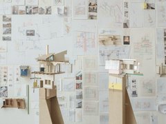 Eine perspektivische Aufnahme eines Ateliers oder Ausstellungsraums, in dem zwei hohe Architekturmodelle auf Sperrholzsockeln vor einer mit Zeichnungen bedeckten Wand stehen. Das linke Modell zeigt eine komplexe, fragmentarische Gebäudestruktur mit Holzkonstruktionen, einem Kamin und einer grünen Dachstütze. Das rechte Modell hat eine rosa Wand und große Fenster. Die gesamte Rückwand ist dicht mit architektonischen Skizzen, Plänen, kleinen Fotos und gefalteten Papiermodellen bedeckt. Die Fülle an Studien vermittelt den Eindruck eines intensiven kreativen Prozesses. Der Boden ist aus dunklem Holz.