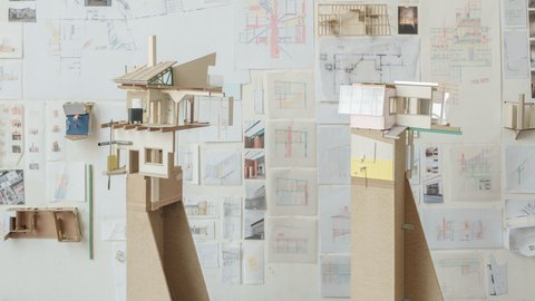 Eine perspektivische Aufnahme eines Ateliers oder Ausstellungsraums, in dem zwei hohe Architekturmodelle auf Sperrholzsockeln vor einer mit Zeichnungen bedeckten Wand stehen. Das linke Modell zeigt eine komplexe, fragmentarische Gebäudestruktur mit Holzkonstruktionen, einem Kamin und einer grünen Dachstütze. Das rechte Modell hat eine rosa Wand und große Fenster. Die gesamte Rückwand ist dicht mit architektonischen Skizzen, Plänen, kleinen Fotos und gefalteten Papiermodellen bedeckt. Die Fülle an Studien vermittelt den Eindruck eines intensiven kreativen Prozesses. Der Boden ist aus dunklem Holz.