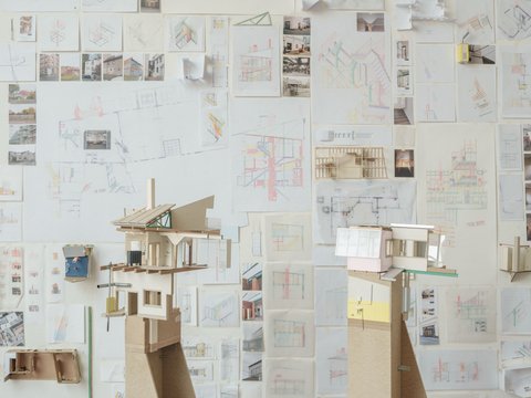 Eine perspektivische Aufnahme eines Ateliers oder Ausstellungsraums, in dem zwei hohe Architekturmodelle auf Sperrholzsockeln vor einer mit Zeichnungen bedeckten Wand stehen. Das linke Modell zeigt eine komplexe, fragmentarische Gebäudestruktur mit Holzkonstruktionen, einem Kamin und einer grünen Dachstütze. Das rechte Modell hat eine rosa Wand und große Fenster. Die gesamte Rückwand ist dicht mit architektonischen Skizzen, Plänen, kleinen Fotos und gefalteten Papiermodellen bedeckt. Die Fülle an Studien vermittelt den Eindruck eines intensiven kreativen Prozesses. Der Boden ist aus dunklem Holz.