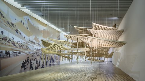"Große weiße Ausstellungshalle mit dynamischer räumlicher Installation. Auf der linken Seite ist eine große projizierte fotografische Komposition mit vielen Personen zu sehen. Gelbe Holz- oder Bambusstrukturen bilden gestaffelte, schwebende Plattformen und Ebenen, die sich durch den Raum ziehen. An den Strukturen sind fotografische Ausdrucke befestigt. Die Installation schafft unterschiedliche Höhenebenen und perspektivische Blickpunkte. Besucher bewegen sich durch diese räumliche Komposition."