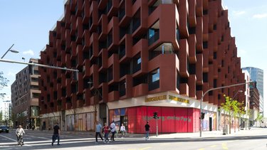 Mehrgeschossiges Gebäude mit rhythmisch gestapelten, rotbraunen Balkonelementen an einer Straßenecke.