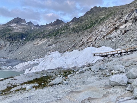 Eine alpine Gebirgslandschaft mit felsigen Hängen und einem Gletscher, der großflächig mit weißen Schutzplanen abgedeckt ist; rechts führt ein hölzerner Steg über das Gestein.