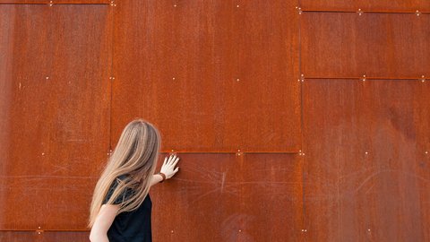 "Rückansicht einer Frau mit langem, graumeliertem Haar in schwarzem Oberteil. Sie steht vor einer großen rostfarbenen, strukturierten Metallplattenwand mit horizontalen Fugen und silbernen Befestigungsschrauben. Die Frau berührt die Wand mit ihrer rechten Hand."