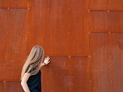 "Rückansicht einer Frau mit langem, graumeliertem Haar in schwarzem Oberteil. Sie steht vor einer großen rostfarbenen, strukturierten Metallplattenwand mit horizontalen Fugen und silbernen Befestigungsschrauben. Die Frau berührt die Wand mit ihrer rechten Hand."