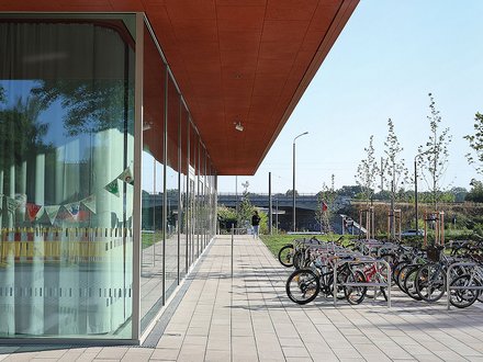 Moderne Architektur mit rotem Vordach über einer breiten Terrasse. Links befindet sich ein Gebäude mit großen Fensterfronten, rechts mehrere Fahrräder in einem Fahrradständer. Die gepflasterte Terrasse führt zu einer grünen Parklandschaft mit jungen Bäumen und blauem Himmel im Hintergrund
