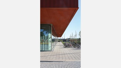 Moderne Architektur mit rotem Vordach über einer breiten Terrasse. Links befindet sich ein Gebäude mit großen Fensterfronten, rechts mehrere Fahrräder in einem Fahrradständer. Die gepflasterte Terrasse führt zu einer grünen Parklandschaft mit jungen Bäumen und blauem Himmel im Hintergrund