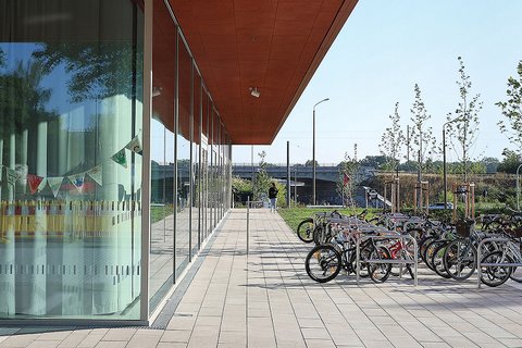 Moderne Architektur mit rotem Vordach über einer breiten Terrasse. Links befindet sich ein Gebäude mit großen Fensterfronten, rechts mehrere Fahrräder in einem Fahrradständer. Die gepflasterte Terrasse führt zu einer grünen Parklandschaft mit jungen Bäumen und blauem Himmel im Hintergrund