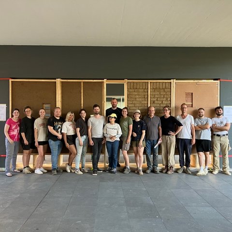 „Gruppenfoto der Workshop-Teilnehmer vor einer großen Musterwand, die verschiedene ökologische Bauweisen mit Holz, Lehmputz und Lehmsteinen zeigt.“