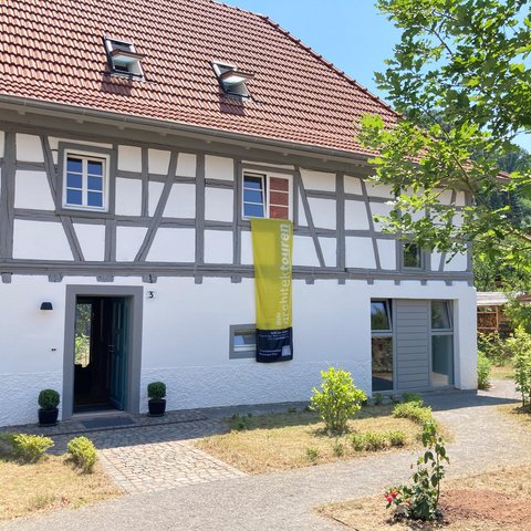 „Ein modernisiertes Fachwerkhaus mit rotem Ziegeldach an einem sonnigen Tag. Aus einem Fenster im Obergeschoss hängt ein langes, gelbes Banner an der weißen Fassade herab.“