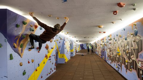 Bouldern im umgestalteten Fußgängertunnel: bunte Klettergriffe an Wänden und Decke, gelbe Zickzack-Linie; Person hängt über Kopf, Gummiplatten-Boden.