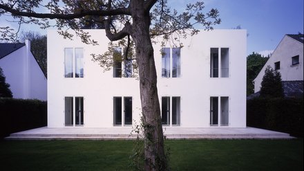 "Zweigeschossiges modernes Wohnhaus mit schneeweißer Fassade. Symmetrisch angeordnete große Fenster in vier Reihen. Breite gepflasterte Terrasse im Vordergrund. Ein großer knorriger Baum mit ausladendem Ast vor der Hausfassade. Grüner Rasen, umgeben von Hecken. Blauer Himmel."