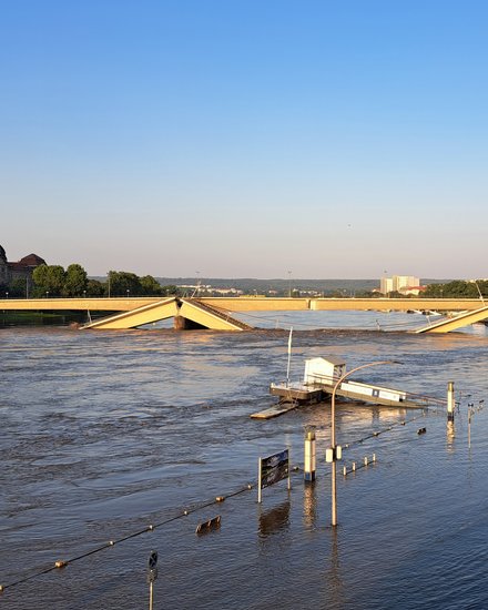 Eine Fotografie zeigt eine teilweise eingestürzte Brücke über einen stark angeschwollenen Fluss, Brückenteile sind abgesackt und ragen schräg ins Wasser, überschwemmte Uferbereiche und versunkene Infrastruktur verdeutlichen die Ausnahmesituation, im Hintergrund ist ein markantes historisches Gebäude sichtbar, wodurch das Bild die Verletzlichkeit technischer Bauwerke zeigt und für die Bedeutung von Instandhaltung, Resilienz und verantwortungsvollem Umgang mit bestehender Infrastruktur steht.